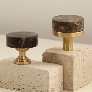 Könnte beinhalten: Zwei dekorative Schrankknöpfe. Jeder Knopf hat eine dunkelbraune Marmorplatte mit goldfarbener Metallbeschlägen. Die Knöpfe sind auf hellbeige, quadratischen Steinblöcken montiert. Die Knöpfe sind ein stilvolles Möbelakzent.