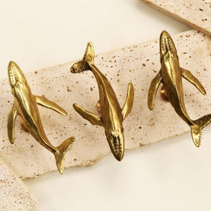 Puede incluir: Tres tiradores de armario dorados con forma de ballena se muestran sobre una superficie clara y texturizada. Los tiradores de ballena están detallados con aletas y colas, y están montados sobre una piedra rectangular.