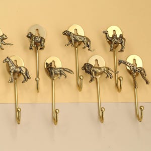 May include: Set of eight brass animal-themed wall hooks. Each hook features a different animal: cheetah, monkey, tiger, camel, horse, wolf, lion, and giraffe. The hooks are mounted on a light yellow surface.