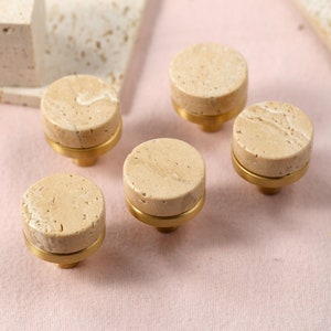 Op de afbeelding: Vijf ronde, beige marmeren kastknoppen met gouden metalen bases. De knoppen zijn in een halve cirkel op een roze oppervlak gerangschikt.