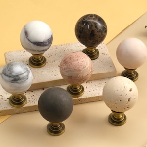 May include: A collection of six spherical cabinet knobs made from various natural stones, including white, gray, brown, pink, and beige marble and travertine. Each knob features a polished stone sphere mounted on a circular antique brass base.