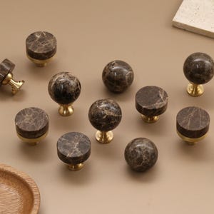 May include: A collection of cabinet knobs featuring dark brown marble tops with white veining and gold-toned metal bases. The set includes both spherical and flat-topped cylindrical shapes, each mounted on a short, sturdy metal post for attachment.