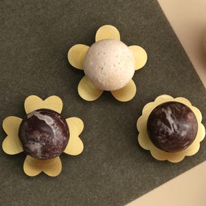 May include: A collection of cabinet knobs featuring spherical stone tops in beige and dark brown with white veining. Each knob is mounted on a gold-toned metal base shaped like a flower with rounded petals, providing a decorative hardware accent.