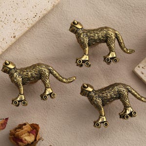 Puede incluir: Tres pomos de armario de metal en tono latón con forma de gatos sobre patines. Cada figura de gato presenta una textura de pelaje detallada y se muestra en posición de pie con cuatro pequeñas ruedas unidas a las patas como tirador.