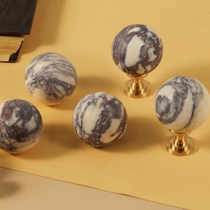 May include: Six marble cabinet knobs with a gold-colored base. The knobs are spherical, with a white and gray marbled pattern. The knobs are arranged on a yellow surface, with a book in the background.