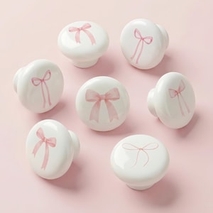 May include: A collection of seven white ceramic cabinet knobs. Each knob features a hand-painted pink bow design. The knobs are arranged on a light pink surface, showcasing their decorative appeal for furniture or home decor projects.