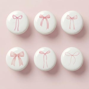 Pode incluir: Seis puxadores decorativos redondos brancos com desenhos de laços rosa. Os laços variam em estilo, alguns com fitas longas e esvoaçantes e outros com um design mais compacto. Os puxadores estão dispostos sobre um fundo rosa claro.