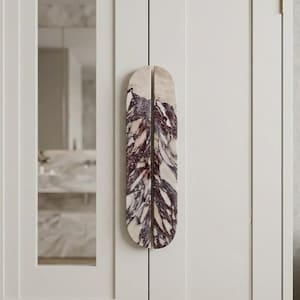May include: A pair of vertical, semi-circular cabinet handles made of polished stone with a white base and deep purple, veined marble patterns. The handles are mounted side-by-side on a light cream-colored cabinet door with a vertical mirror panel.