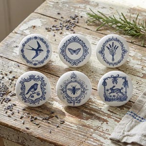 Op de afbeelding: Zes witte keramische kastknoppen met blauwe illustraties van een vogel, vlinder, bij, konijn en lavendel. De knoppen liggen op een verweerd houten oppervlak, met rozemarijn en lavendel.