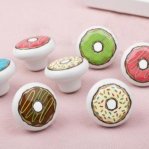 May include: A collection of seven white ceramic cabinet knobs, each adorned with a colorful donut design. The donuts feature various toppings, including pink, green, brown, and blue frosting, with sprinkles and icing details. These knobs are ideal for adding a playful touch to furniture.
