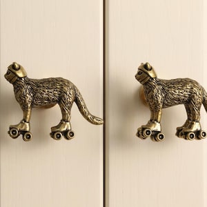 May include: Two antique brass-toned cabinet knobs shaped like cats wearing helmets and roller skates. Each cat figure is depicted in a standing profile position with textured fur details, attached to a circular base for mounting on cabinet doors.