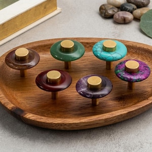 May include: A collection of six decorative cabinet knobs with brass accents, displayed on a wooden oval tray. The knobs feature various colors and patterns, including brown, green, turquoise, and purple. The knobs are designed for furniture and home decor.