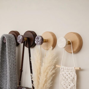 Könnte beinhalten: Eine Sammlung von Wandhaken aus Holz mit dekorativen Knöpfen. Die Haken halten einen grauen Schal, eine braune Ledertasche, getrocknete Pflanzen und einen Makramee-Wandbehang. Die Haken haben eine natürliche Holzoberfläche und Marmorakzente.