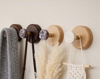May include: A collection of wooden wall hooks with decorative knobs. The hooks are holding a gray scarf, a brown leather bag, dried plants, and a macrame wall hanging. The hooks have a natural wood finish and marble accents.