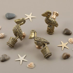 Könnte beinhalten: Bronzefarbene Schrankknöpfe in Form von Pelikanen. Jeder Knopf zeigt einen detaillierten Pelikan, der auf einem Holzpfosten sitzt. Die Knöpfe sind von Muscheln, Seesternen und kleinen Steinen umgeben, was ein Küstenmotiv ergibt.