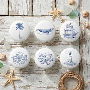Può includere: Un set di sei pomelli per mobili in ceramica bianca con disegni nautici blu: palma, balena, veliero, conchiglie, polpo e faro. Disposti su una superficie di legno bianco invecchiato, con stelle marine e conchiglie.