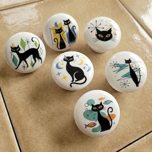Puede incluir: Un conjunto de seis pomos de cerámica blanca. Cada pomo presenta un diseño de gato negro con varios acentos de estilo moderno de mediados de siglo, incluyendo formas geométricas, estrellas y peces. Fondo neutro.