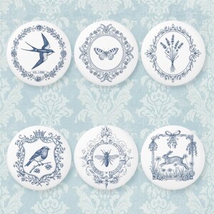 Könnte beinhalten: Set aus sieben weißen Keramik-Schrankknöpfen mit blauen Illustrationen. Designs umfassen eine Schwalbe, einen Schmetterling, Lavendel, einen Vogel, eine Biene und ein Kaninchen. Jeder Knopf hat ein anderes Design, ideal für einen Hauch von Eleganz in jedem Raum.