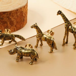 Pode incluir: Conjunto de quatro puxadores de gaveta de animais em tom dourado. Os puxadores representam uma girafa, um cavalo, um rinoceronte e uma raposa.