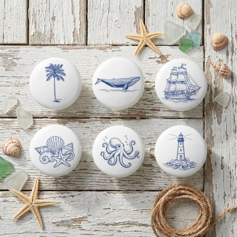 以下が含まれることがあります： 白いセラミック製のキャビネットノブ6個セット。青い航海デザインで、ヤシの木、クジラ、帆船、貝殻、タコ、灯台が描かれています。古びた白い木製の表面に配置され、ヒトデや貝殻が散らばっています。