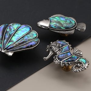 May include: Decorative cabinet knobs in the shape of a seashell, fish, and seahorse. The knobs have a silver finish and iridescent abalone shell inlays, with a brass-colored base. The seashell knob is fan-shaped.
