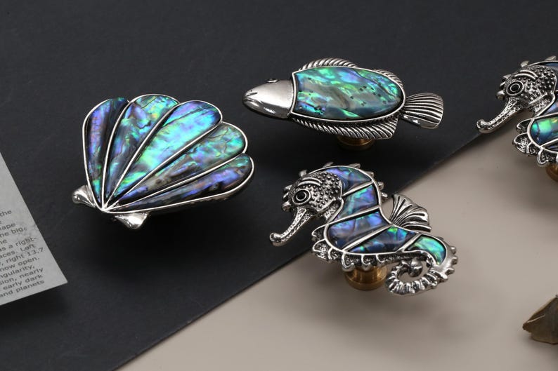 May include: Decorative cabinet knobs in the shape of a seashell, fish, and seahorse. The knobs have a silver finish and iridescent abalone shell inlays, with a brass-colored base. The seashell knob is fan-shaped.