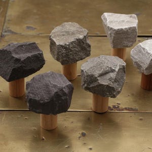 Può includere: Sei pomelli decorativi per mobili. Ogni pomello presenta una parte superiore in pietra naturale nei toni del grigio e nero, montata su un tassello di legno. I pomelli sono disposti su una superficie metallica, mostrando il loro design rustico.