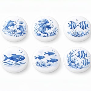 Op de afbeelding: Set van zes witte keramische kastknoppen met blauwe aquarel visontwerpen. Elke knop heeft een andere aquatische scène, waaronder vissen, koraal en zeewier. De knoppen zijn rond en hebben een glanzende afwerking.