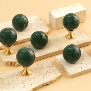 May include: Six green marble cabinet knobs with gold bases on a light beige stone surface.