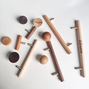 Op de afbeelding: Een verzameling houten kastknoppen en -grepen in verschillende vormen en bruintinten. Sommige stukken hebben messing hardware. Het woord "Allhome" is op twee van de grepen gedrukt. De knoppen en grepen zijn gerangschikt op een wit oppervlak.