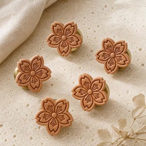 以下が含まれることがあります： 5つの花びらを持つ桜の形をした引き出し用ノブ5個セット。各ノブは、マット仕上げのライトブラウンのフラワーモチーフで、花びらのエンボス加工と中央の円形のおしべが特徴です。円形のゴールドトーンの金属製ベースに取り付けられています。
