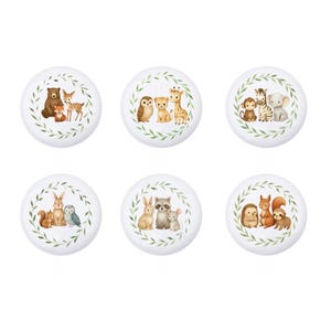 May include: Six white ceramic cabinet knobs with watercolor illustrations of woodland and safari animals. Each knob features a different group of animals, including a bear, deer, fox, owl, giraffe, zebra, elephant, and more, surrounded by a green leaf wreath.