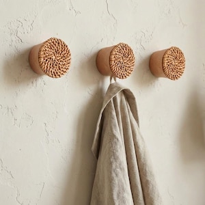 May include: Three round wall hooks with a natural wood base and woven rattan front. One hook holds a beige linen garment. The hooks are mounted on a textured, off-white wall, offering a rustic, minimalist aesthetic.
