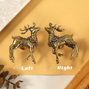 May include: Two antique brass deer-shaped cabinet knobs. The knobs are detailed with antlers and fur, and are labeled "Left" and "Right". The background is a warm yellow with a wooden and marble accent.