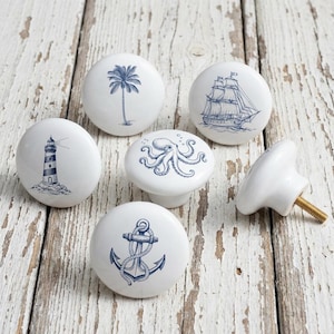 以下が含まれることがあります： 白のセラミック製キャビネットノブのコレクションで、航海をテーマにしたデザインです。各ノブには、灯台、帆船、ヤシの木、錨、タコなど、さまざまな青色のイラストが描かれています。1つのノブはネジ付きで表示されています。