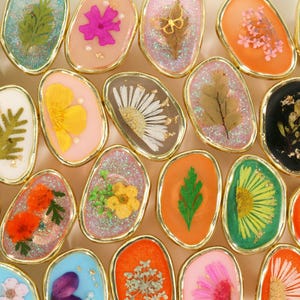 Puede incluir: Una colección de anillos ovalados de resina con bordes dorados. Cada anillo presenta una flor o hoja seca diferente encapsulada en resina transparente, con colores como rosa, naranja, verde y amarillo.