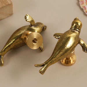 Puede incluir: Dos tiradores de armario con forma de foca, color latón. Los tiradores presentan detalles de aletas y aletas caudales. Diseñados para ser montados en un armario o cajón. El fondo incluye un bloque de madera y una concha.