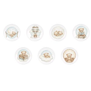 Könnte beinhalten: Set aus sieben runden dekorativen Schrankknöpfen mit Aquarellillustrationen von Teddybären. Jeder Knopf zeigt eine andere Szene, darunter einen Bären in einer Hängematte, einen Heißluftballon und einen Bären, der ein Buch liest. Die Knöpfe sind hauptsächlich weiß mit sanften Pastellfarben.