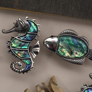 May include: Decorative seahorse and fish-shaped cabinet knobs. The seahorse and fish are silver-toned metal with iridescent abalone shell inlays of green, blue, and teal. The seahorse has a curled tail. The fish has a rounded body and a fin.