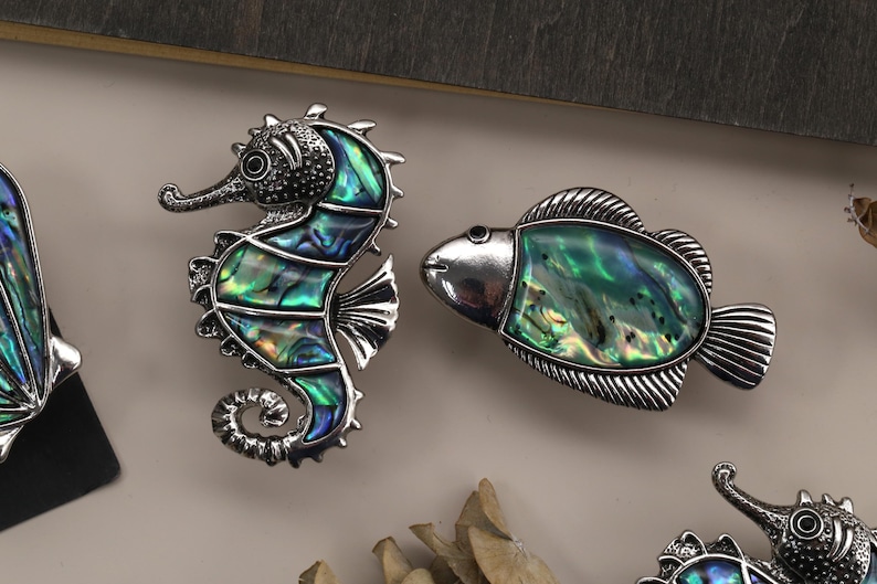 May include: Decorative seahorse and fish-shaped cabinet knobs. The seahorse and fish are silver-toned metal with iridescent abalone shell inlays of green, blue, and teal. The seahorse has a curled tail. The fish has a rounded body and a fin.