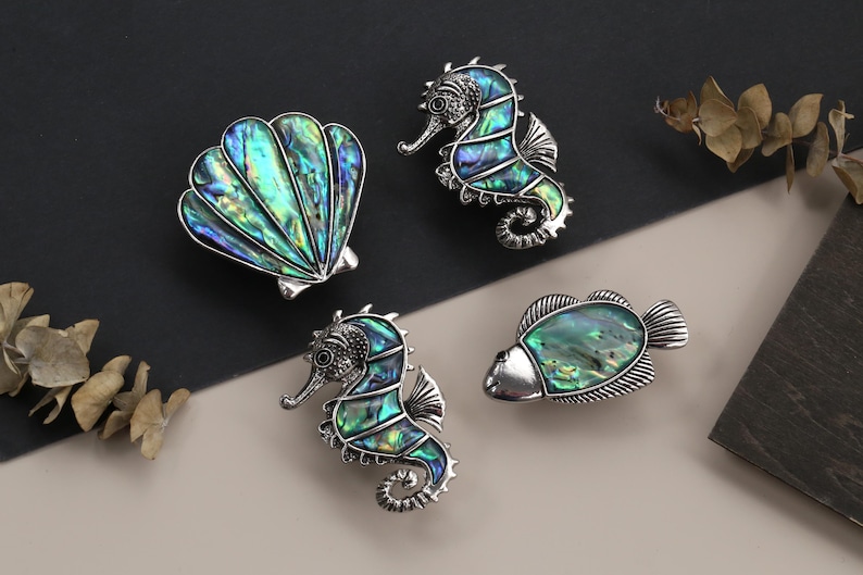 May include: A collection of ocean-themed jewelry pieces, including a seashell, two seahorses, and a fish, all with iridescent abalone shell inlays and silver-toned metalwork. The pieces are displayed on a neutral background.