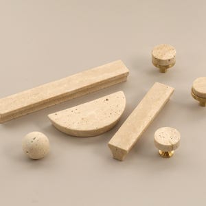 Peut inclure: Ensemble de quincaillerie de meubles en pierre beige, comprenant des boutons, des poignées et un élément sphérique. La quincaillerie a une texture tachetée et est posée sur un fond neutre. Les boutons ont des bases en laiton.