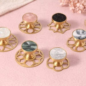 Może przedstawiać: Kolekcja dekoracyjnych gałek do szafek ze złotymi podstawami i różnymi kamiennymi lub marmurowymi blatami. Gałki mają różne wzory i kolory, w tym biały, różowy, czarny i zielony. Gałki są ułożone na różowej powierzchni.