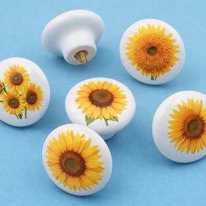 Op de afbeelding: Witte keramische kastknoppen met een zonnebloemontwerp. Elke knop heeft een heldergele zonnebloem met een bruin hart op een witte achtergrond. De knoppen staan tegen een lichtblauwe achtergrond.