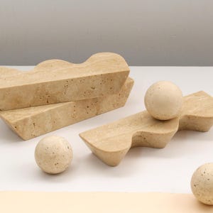 Pode incluir: Uma coleção de objetos de pedra travertino bege, incluindo três blocos retangulares com bordas superiores onduladas e três esferas lisas. A pedra natural apresenta uma superfície porosa e texturizada com variações de cor creme.