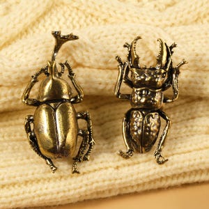 以下が含まれることがあります： 真鍮色のアンティークなカブトムシのブローチ2つ。ブローチは足、触角、質感のあるボディが詳細に表現されています。クリーム色のニットの背景に配置されています。