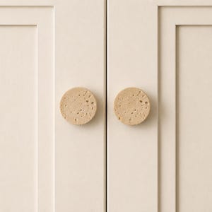 May include: Two circular cabinet knobs made of light beige, porous stone with a natural, pitted texture. The knobs are mounted on a pair of off-white cabinet doors featuring a simple, clean-lined, recessed panel design and a vertical seam.
