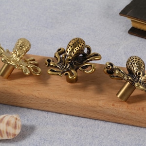 Może przedstawiać: Trzy mosiężne gałki do szafek w kształcie ośmiornicy, prezentowane na drewnianym bloku. Gałki mają szczegółowe macki i teksturowany korpus. Wykończenie jest w ciepłym, antycznym złotym kolorze.