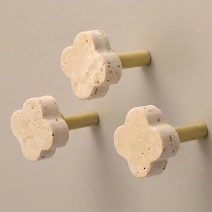 Op de afbeelding: Drie kastknoppen in de vorm van een klavertjevier, gemaakt van beige, poreuze steen met een getextureerd oppervlak. Elke knop is bevestigd aan een cilindrische, mat goudkleurige metalen basis die naar buiten steekt vanuit het midden.