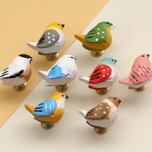 Op de afbeelding: Een verzameling van acht kleurrijke, handgeschilderde houten vogelknoppen. Elke vogel heeft een uniek ontwerp met verschillende kleuren zoals geel, blauw, groen, rood en roze. De knoppen zijn gemonteerd op goudkleurige bases.
